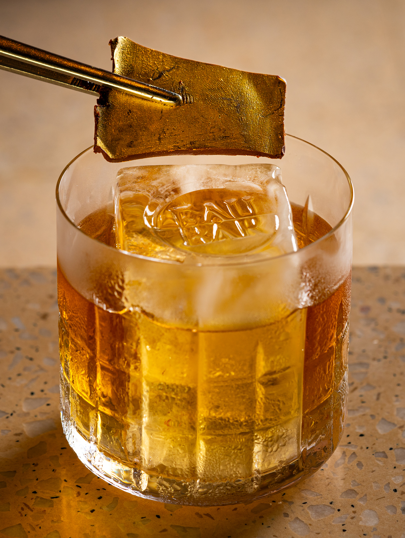 A gold-coloured cocktail in a tumbler with an ice cube featuring the inscription, 'Eau Bar'